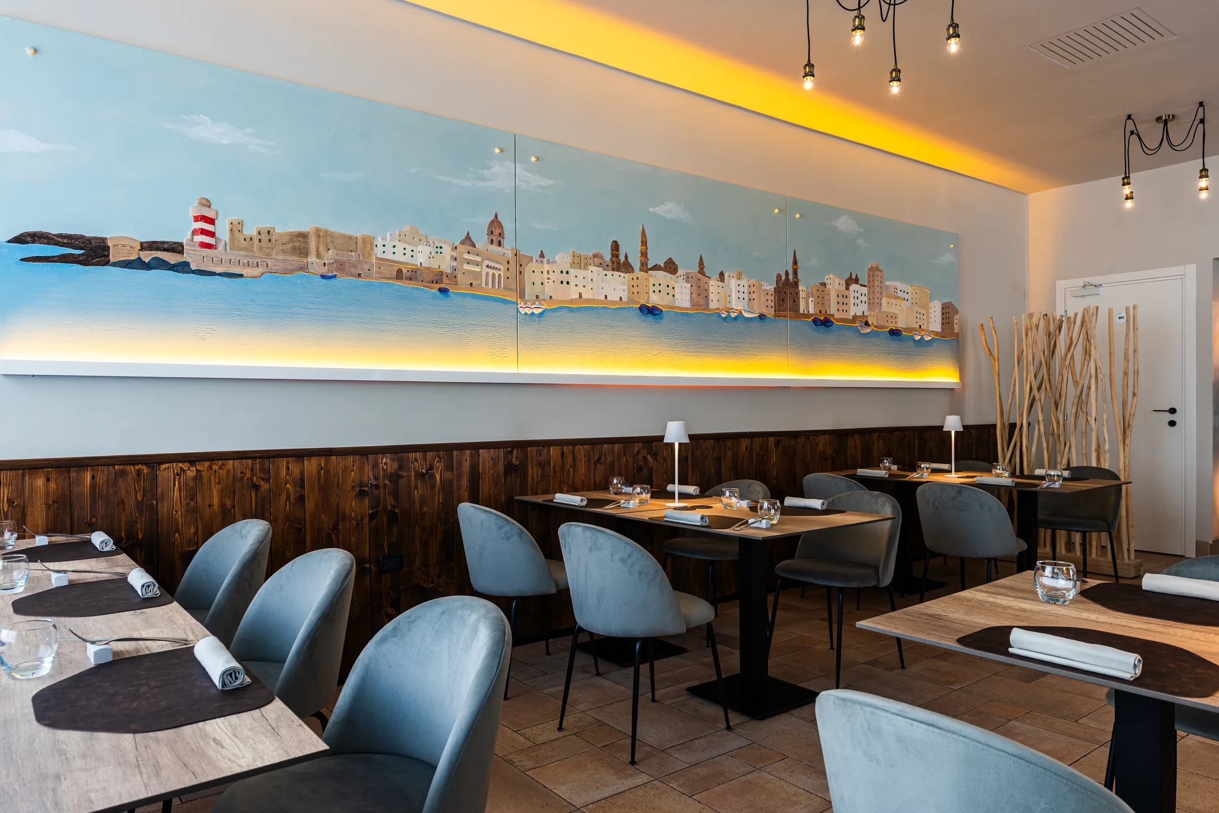 Sala di DDO JÈ con il murales dello skyline di Monopoli e tavoli apparecchiati.