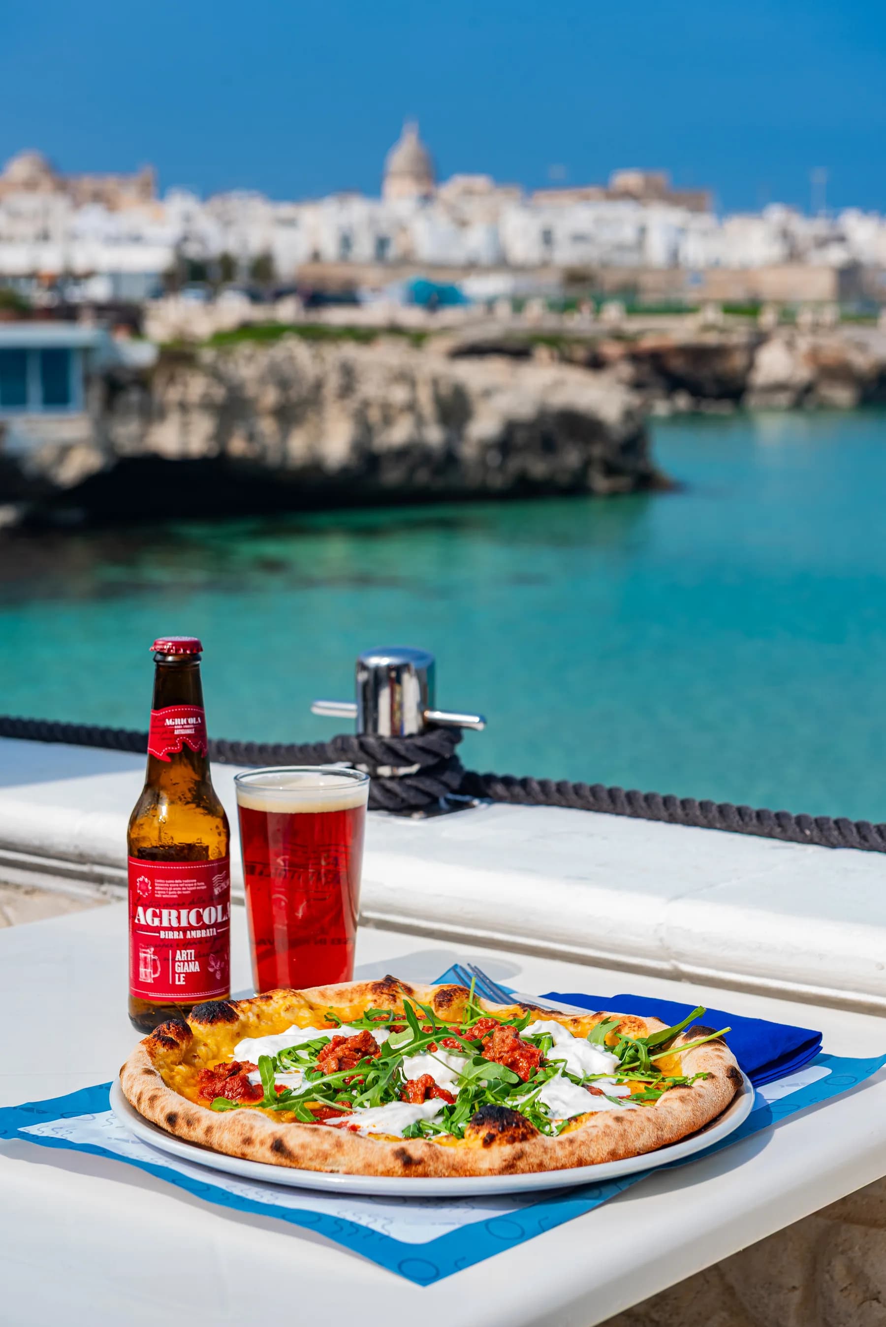 Pizza e birra su un tavolo sul mare con Monopoli sullo sfondo.
